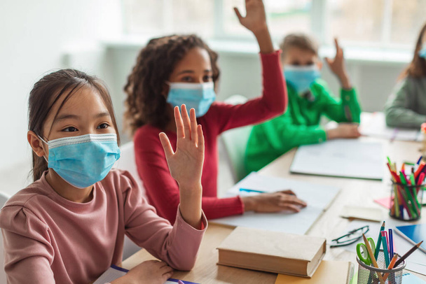 Diverse School Children Wearing Face Masks Raising Arms Having Class Sitting At Desk In Modern Classroom At School. Education During Coronavirus Pandemic. Selective Focus - Фото, изображение