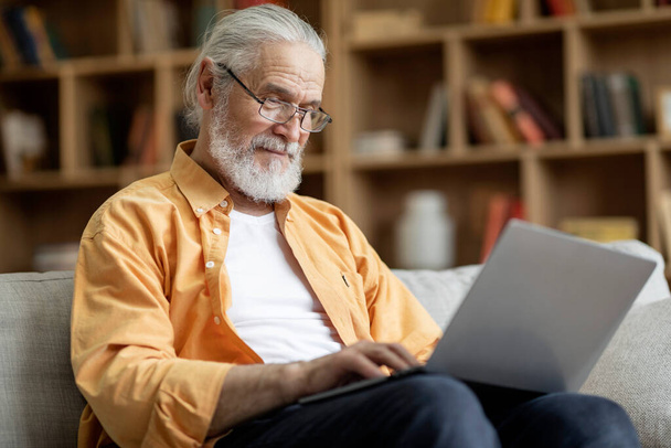 Old man in eyeglassses using modern laptop at home, handsome grandfather sitting on sofa with computer on his lap in living room, typing on notebook keyboard, sending emails, copy space - 写真・画像