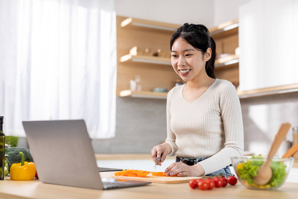 Cheerful beautiful millennial chinese woman cooking dinner at home, looking for nice recipe on Internet, watching food vlog, using laptop, kitchen interior, copy space. Cooking, culinary - Photo, image