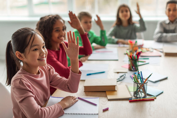 Happy Multiethnic School Kids Learning Raising Arms During Lesson Sitting At Desk In Classroom At School. Modern Education And Knowledge Concept. Selective Focus - Foto, Bild
