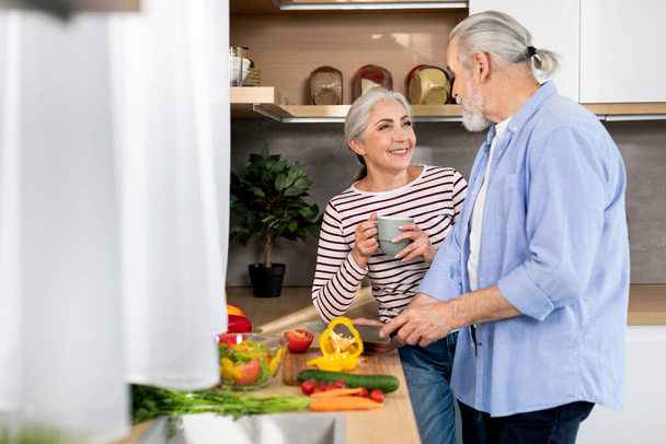 Portrait of beautiful senior couple preparing lunch in kitchen at home, happy elderly man cooking healthy food for his smiling wife, making fresh vegetable salad, cheerful lady drinking tea - Photo, image