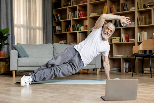 Healthy handsome bearded senior man exercising on yoga mat at home, wearing sportswear, watching fitness video, attending online workout, cozy living room interior, copy space, full length - Fotoğraf, Görsel