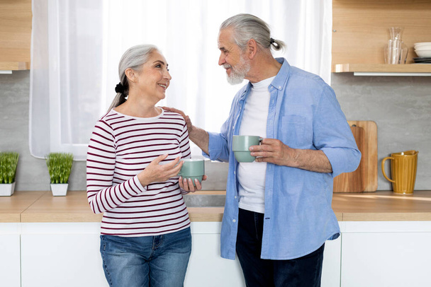 Happy Senior Couple Drinking Morning Coffee And Chatting Together In Kitchen At Home, Loving Elderly Spouses Enjoying Retirement Time, Old Man And Woman Looking At Each Other And Smiling, Copy Space - Фото, зображення