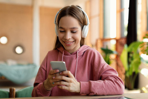 Happy teenager female student in headphones typing message on cellphone, chatting with friends, sitting at home. Remote communicate, modern technology concept - Foto, Imagem
