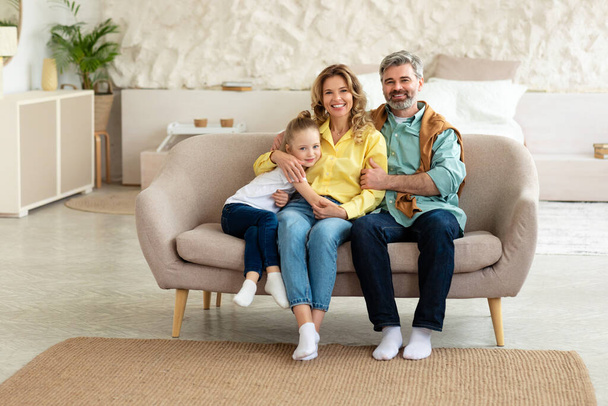 Cheerful Family Of Three Sitting On Couch In Modern Living Room Indoor. Happy Middle Aged Parents Hugging Daughter Kid Smiling To Camera Posing Together At Home. Parenthood Happiness - Фото, изображение
