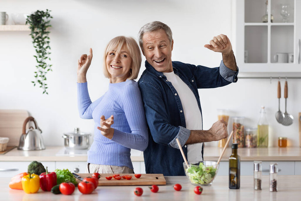 Funny senior husband and wife having fun while cooking at kitchen, positive elderly man and woman in casual making healthy salad and dancing, cheerfully smiling at camera, copy space - Zdjęcie, obraz