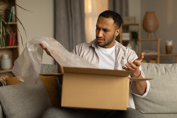 Dissatisfied black male customer opening box from online store, taking out packaging, unhappy with received item after unboxing parcel at home. Remote shopping, wrong delivery concept - Foto, Imagen