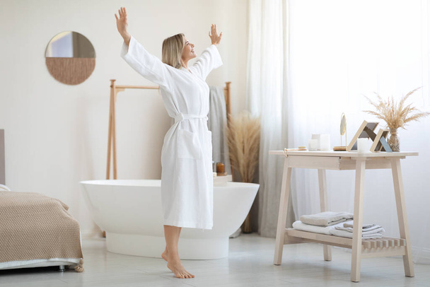 Full length shot of happy beautiful blonde woman in white bathrobe enjoying sunny morning, dancing by white modern open space bedroom, raising hands up and smiling, copy space - Photo, image