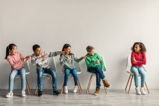 Bullying At School. Preteen Children Laughing At Victimized Black Schoolgirl Sitting Over Gray Wall In Modern Classroom Indoor. Kids And Social Problems Concept. Full Length - 写真・画像