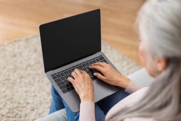 Cropped caucasian elderly gray-haired woman typing on laptop with blank screen in living room interior. Work remotely, business online, good website and technology at home during covid-19 quarantine - Foto, Imagen