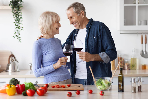 Romantic senior spouses drinking red wine while cooking together at home, loving elderly husband and wife preparing dinner, having conversation, kitchen interior, copy space. Marriage, relationships - Photo, Image
