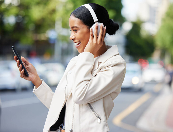 Happy city girl with phone and headphones on video call while walking relax in the street to work or home. Black woman or student laughing at joke, meme or online video while on travel to a location. - Fotografie, Obrázek
