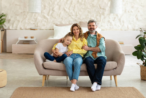 Happy Parents Posing With Cute Little Daughter, Hugging Kid Smiling To Camera Sitting On Sofa At Home. Full Length Shot Of Loving Family Of Three Posing In Modern Living Room Indoor - Fotografie, Obrázek