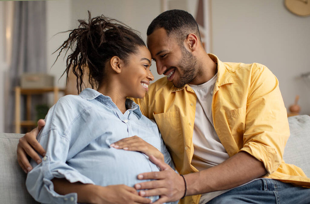 Pregnancy And Childbirth. Loving Black Couple Hugging Expecting Baby, Husband Touching Pregnant Wifes Belly Sitting On Couch At Home. Family Enjoying Expectation. Happiness And Love - Фото, изображение