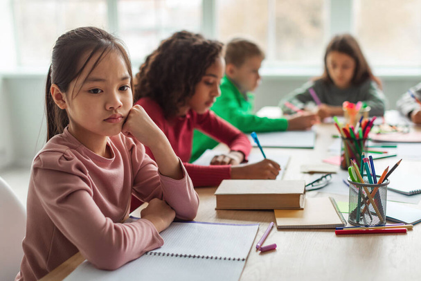 Unhappy Asian Schoolgirl Thinking Having Problem With Education Sitting While Her Diverse Classmates Learning Lesson In Modern Classroom At School. Educational Issues Concept. Selective Focus - 写真・画像