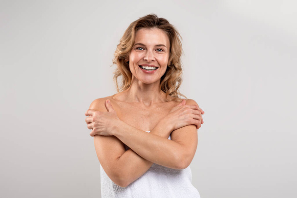 Enjoying Beauty. Happy Attractive Middle Aged Lady Wrapped In Bath Towel Embracing Herself, Beautiful Cheerful Female Smiling At Camera While Posing Over Light Grey Studio Background, Copy Space - Photo, image