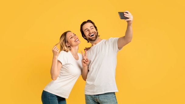 Cheerful excited young caucasian couple in white t-shirts have fun, make peace sign and selfie on phone, isolated on yellow background, panorama. Love, relationship photo for blog and social networks - Foto, Imagen