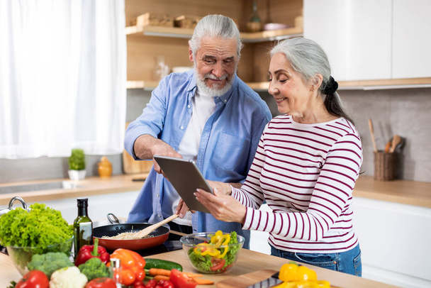 Online Recipe. Happy Senior Couple Using Digital Tablet In Kitchen While Cooking Dinner Together, Smiling Elderly Spouses Preparing Tasty Food At Home And Browsing Internet On Modern Gadget - Photo, image