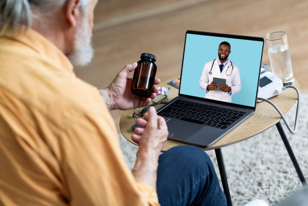 Unrecognizable elderly man seating on sofa, making distant video call, looking at laptop screen, communicating with black doctor therapist online, asking how to take medicine. Telemedicine concept - Foto, afbeelding