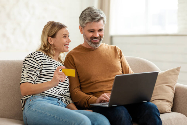 Online Shopping. Happy Middle Aged Couple Making Internet Payment Using Laptop Computer And Credit Card Sitting On Sofa At Home. Ecommerce, Technology And Consumerism - Φωτογραφία, εικόνα