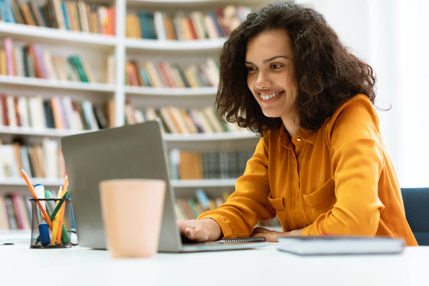 Happy mixed race lady sitting at desk, using laptop and watching tutorial, lecture or webinar, studying online in library interior, looking at screen and smiling - Photo, Image