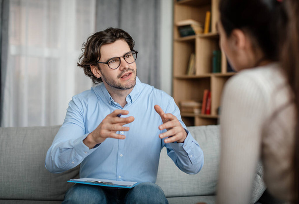Emotional young european guy doctor speaks and gesticulates with woman patient in office clinic interior. Medical support, psychological therapy, session with professional, mental help and treatment - Foto, imagen