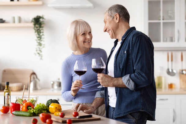 Positive elderly spouses drinking red wine while preparing meal at home, cutting fresh vegetables, holding glasses with drink, having conversation, enjoying time together, kitchen interior, copy space - Fotografie, Obrázek