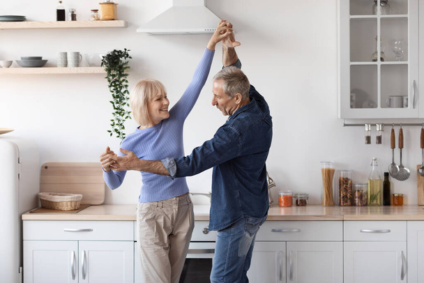 Happy senior couple having fun at home, cheerful elderly woman dancing with her husband, looking at each other and laughing, holding hands, white cozy kitchen interior, copy space - Photo, Image