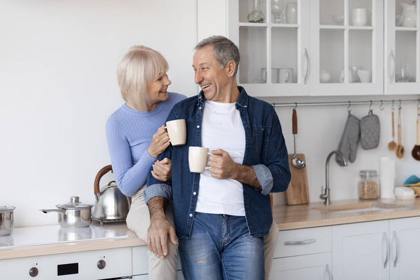 Cheerful elderly spouses enjoying time together at home, drinking coffee, happy senior lady sitting on kitchen table, hugging her handsome husband, couple having conversation, copy space - Fotó, kép