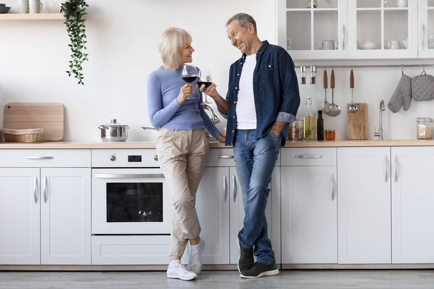 Happy senior couple drinking red wine at kitchen, elderly man and woman in casual outfits standing by kitchen table with glasses, having conversation, enjoying time together, copy space, full length - Photo, Image