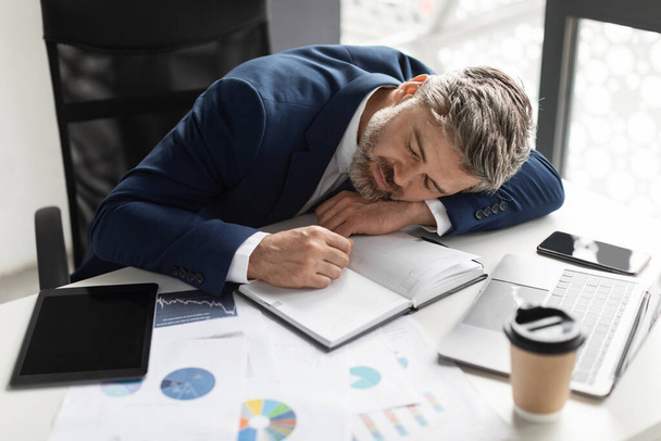 Overworked Middle Aged Businessman Napping At Workplace In Office, Mature Male Entrepreneur In Suit Sleeping At Desk With Financial Reports And Coffee, Tired After Long Working Day, Above View - Photo, image