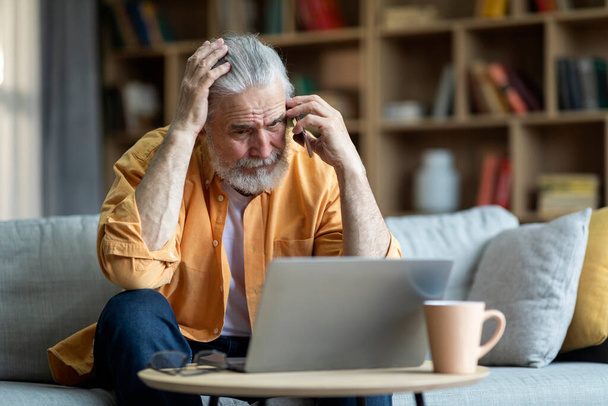 Upset handsome elderly man with beard businessman sitting on couch, using modern laptop and having phone conversation with assistant, touching his head, working from home, having problems, copy space - Fotoğraf, Görsel