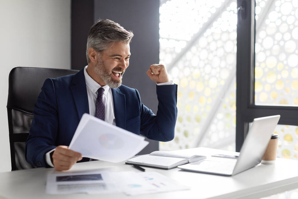 Business Success. Portrait Of Overjoyed Middle Aged Businessman With Documents And Laptop In Office, Happy Male Entrepreneur Celebrating Successful Deal, Shaking Fists And Exclaining With Joy - Фото, зображення