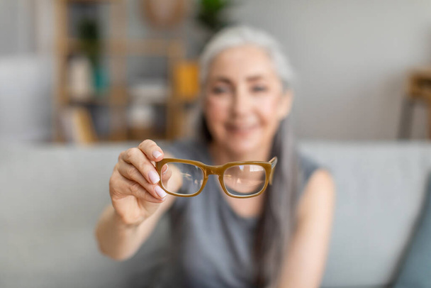 Cheerful caucasian elderly gray-haired female holding glasses in living room interior, selective focus, blurry. Health care, poor eyesight, age-related problems, myopia and advertising, free space - Photo, image