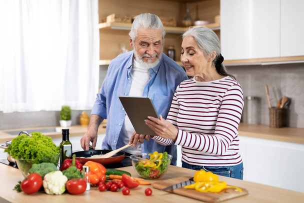 Happy Elderly Spouses Cooking Food In Kitchen And Using Digital Tablet Together, Married Senior Couple Checking Online Recipe While Preparing Healthy Vegetable Meal For Lunch At Home, Copy Space - Valokuva, kuva