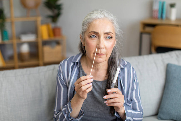 Concentrated caucasian old gray-haired lady doing covid-19 test by herself in living room interior. Health care at home, medicine, fight against epidemic, first symptoms of disease and diagnostic - Foto, Imagem
