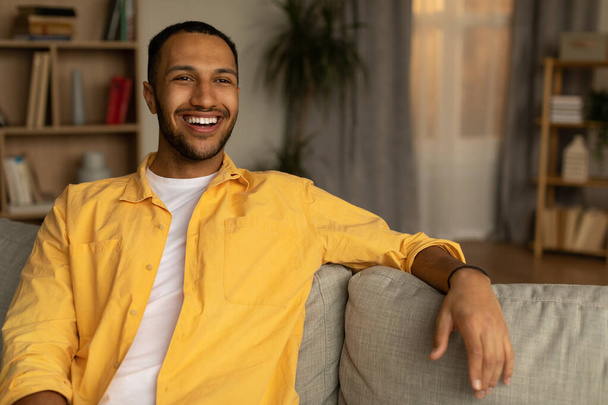 Portrait of attractive young black guy smiling, sitting on couch at home, empty space. Millennial African American man showing positive emotions, expressing joy and happiness - Photo, Image