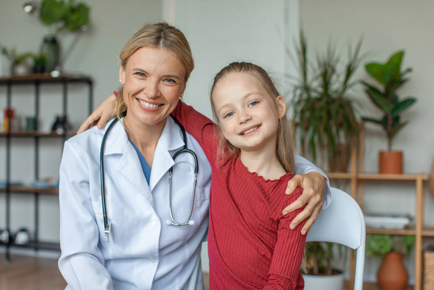 Portrait of friendly female doctor embracing with little cute girl patient in hospital, looking and smiling togethe at camera. Good children healthcare concept - Foto, Imagen