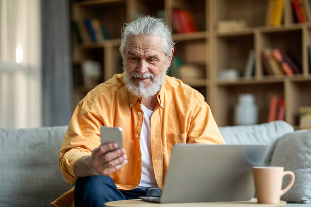 Happy senior businessman working from home, sitting on couch, using modern laptop and cell phone, looking at smartphone screen and smiling, enjoying business mobile app, copy space - Photo, image