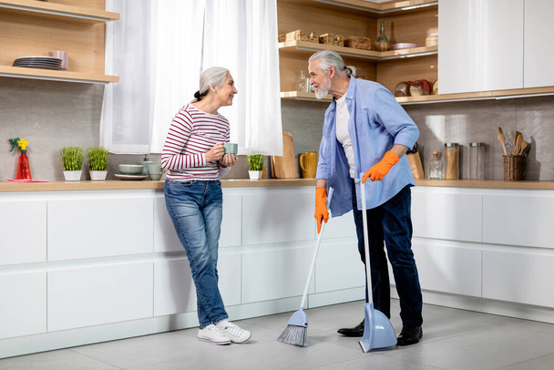 Portrait of happy senior spouses making cleaning in kitchen together, smiling elderly couple sharing domestic chores, woman drinking tea while her husband mopping floor at home, copy space - Фото, изображение