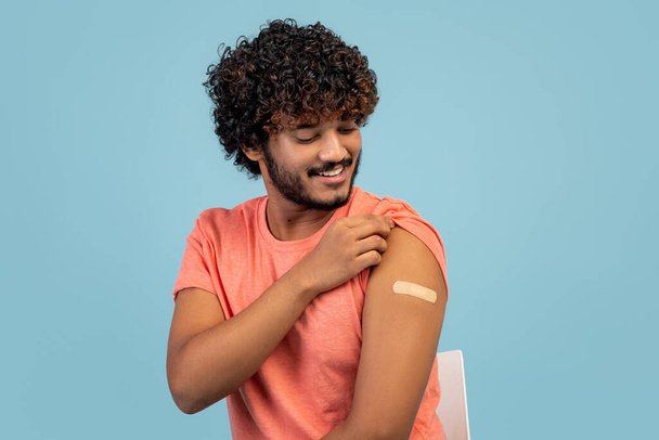 Happy curly young indian guy sitting on chair over blue studio background, looking at medical band on his shoulder and smiling, got vaccinated against flu or COVID-19, copy space, closeup - Foto, imagen