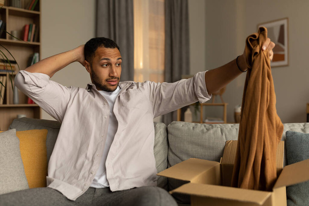 Dissatisfied black male client opening box from online shop and looking at wrong delivered clothes, unhappy with received item, feeling confused after unboxing package at home - Foto, Imagen