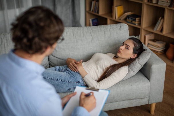 Sad millennial european woman suffering from mental problems lies on couch, talk with man doctor in clinic interior. Therapy, session with professional, help and prescription for depression treatment - Foto, Bild