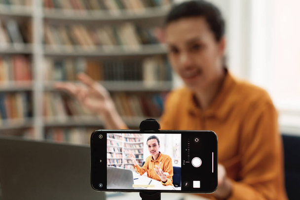 Online studying, virtual class. Mixed race female teacher shooting lesson on smartphone camera, sitting in library and talking to cellphone, selective focus - Zdjęcie, obraz