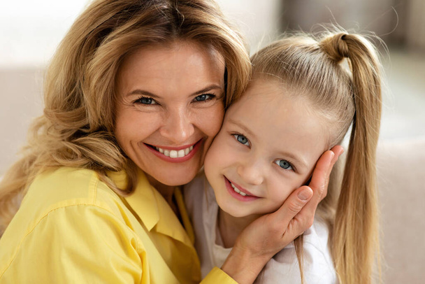 Portrait Of Loving Middle Aged Mother And Little Daughter Embracing Smiling To Camera Indoor. Mommy And Her Kid Posing Together At Home. Mothers Day, Family Lifestyle Concept - Foto, Imagen