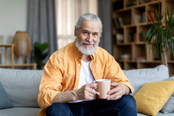 Cheerful attractive grandfather sitting on couch, holding mug, drinking herbal tea while chilling alone at home, looking at camera and smiling, living room interior, copy space - 写真・画像