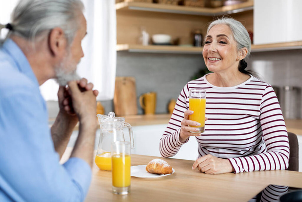 Happy Senior Woman Drinking Orange Juice While Having Breakfast With Husband In Kitchen, Cheerful Elderly Spouses Enjoying Morning Meal And Chatting With Each Other, Closeup, Selective Focus On Lady - Photo, image