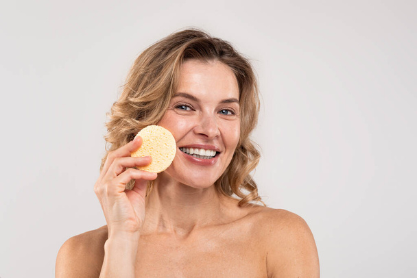Beauty Care. Attractive Middle Aged Woman Holding Cosmetic Sponge And Smiling At Camera, Beautiful Mature Lady With Bare Shoulders Cleansing Face From Makeup Standing Over Light Studio Background - Foto, Imagen