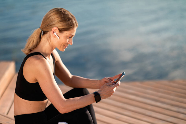 Smiling Middle Aged Woman Relaxing With Smartphone After Outdoor Training, Beautiful Sporty Female Sitting On Wooden Pier Near River Using Mobile Phone For Messaging With Friends, Copy Space - 写真・画像