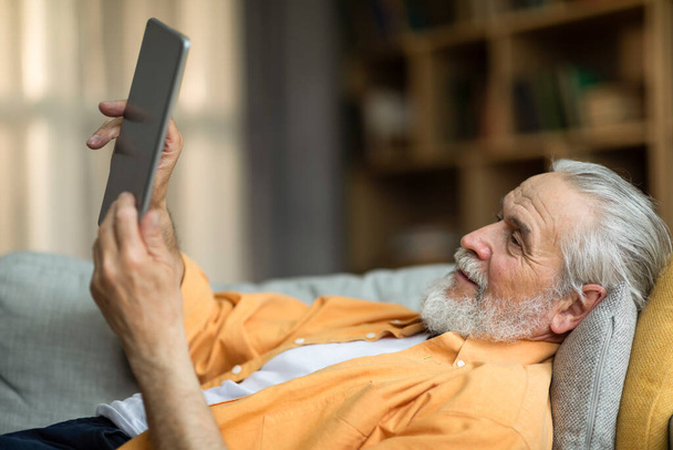 Positive attractive senior man reclining on couch at home, using modern digital tablet, checking social media while chilling alone at weekend, looking at screen and smiling. Leisure and gadgets - 写真・画像
